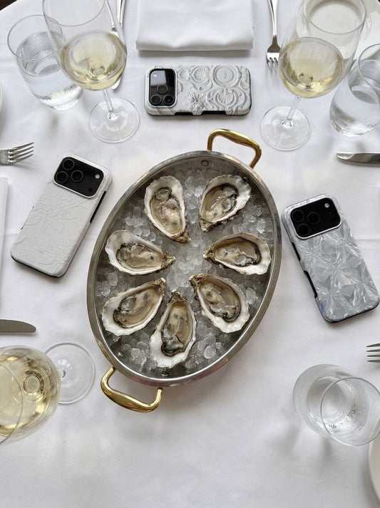 A stylish table setting with white wine glasses, oysters on ice in a silver dish, and trendy phone cases in flerfarvet, hvid, and beige tones.