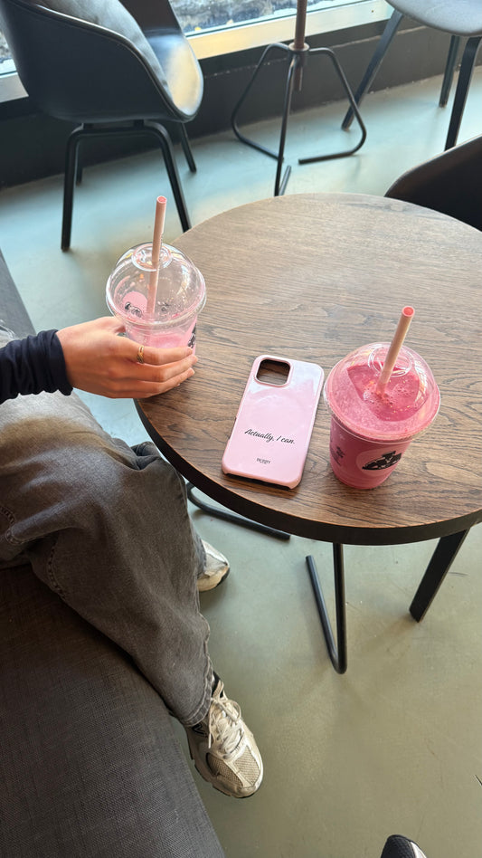 Pink phone case with "Actually, I can" text on a wooden table next to two pink smoothies in a trendy café setting with a casually dressed person nearby.