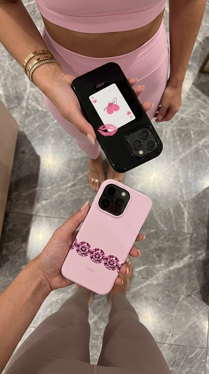 Two hands holding trendy phone cases, one black with a pink diamond-themed card and one pink with a rosaguld floral pattern, styled with feminine outfits.