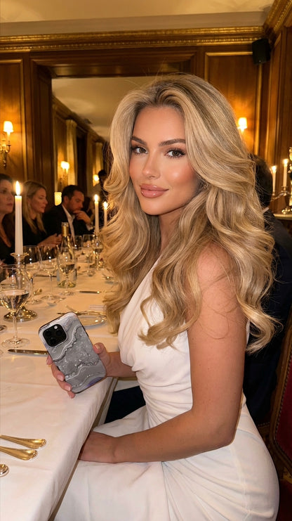 Trendy woman with voluminous blonde curls at elegant dinner holding a sleek silver marble quartz phone case with multicolored gray and silver pattern.