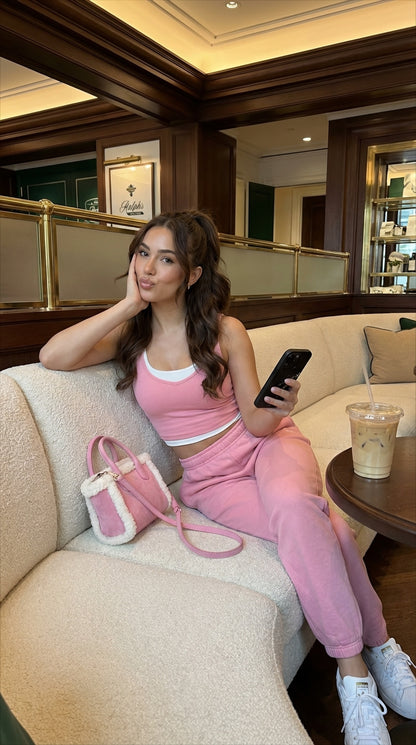 Trendy girl relaxing on a plush sofa in pink sporty outfit holding a phone with a stylish pink and gold multicolored bag nearby and iced coffee on the table.
