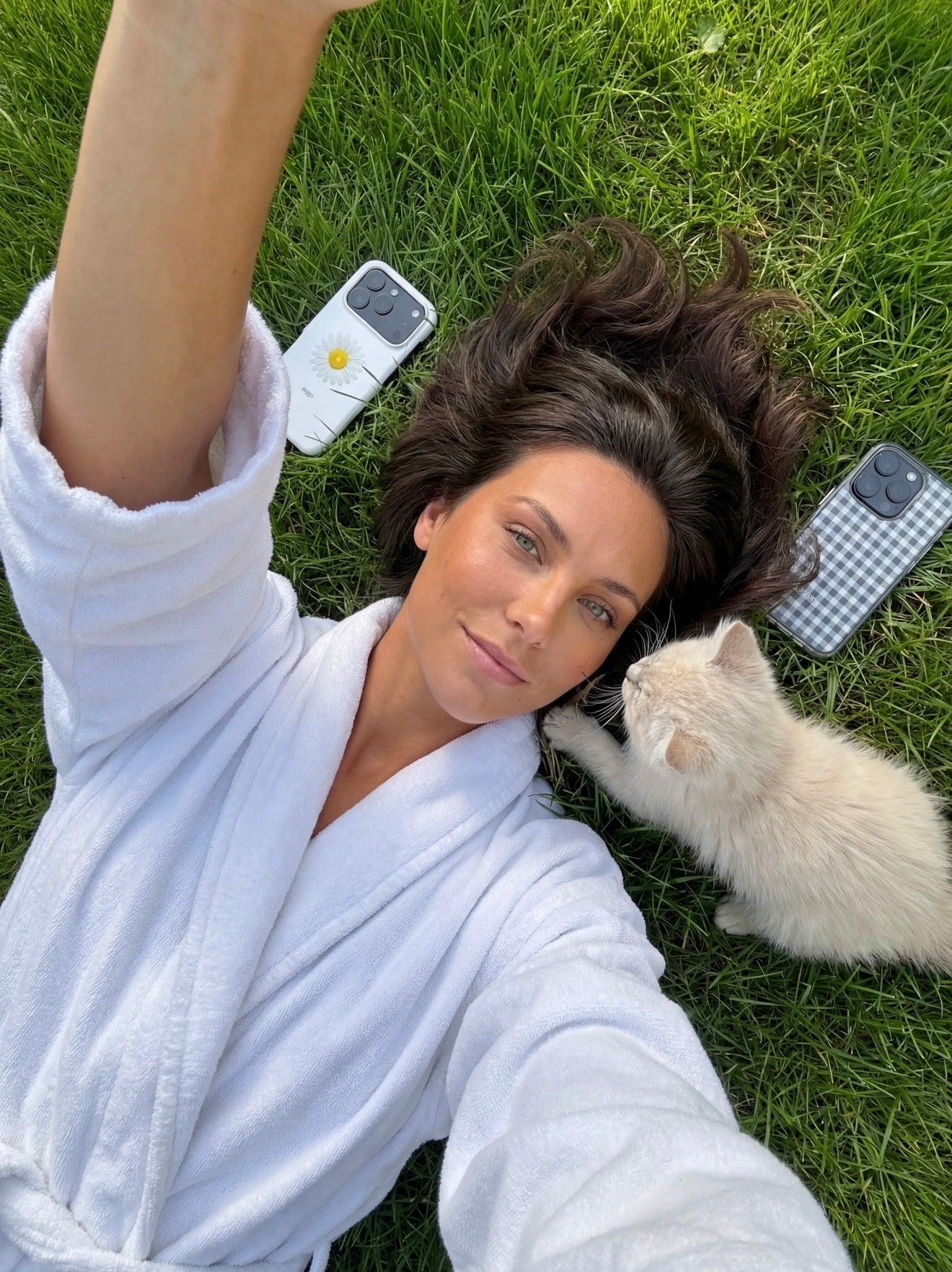 Trendy woman in a white robe lying on grass with a small cat, featuring two phones with Klar and Hvid color-patterned cases beside her.