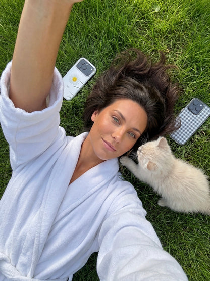 Trendy woman in a white robe lying on grass with a small cat, featuring two phones with Klar and Hvid color-patterned cases beside her.