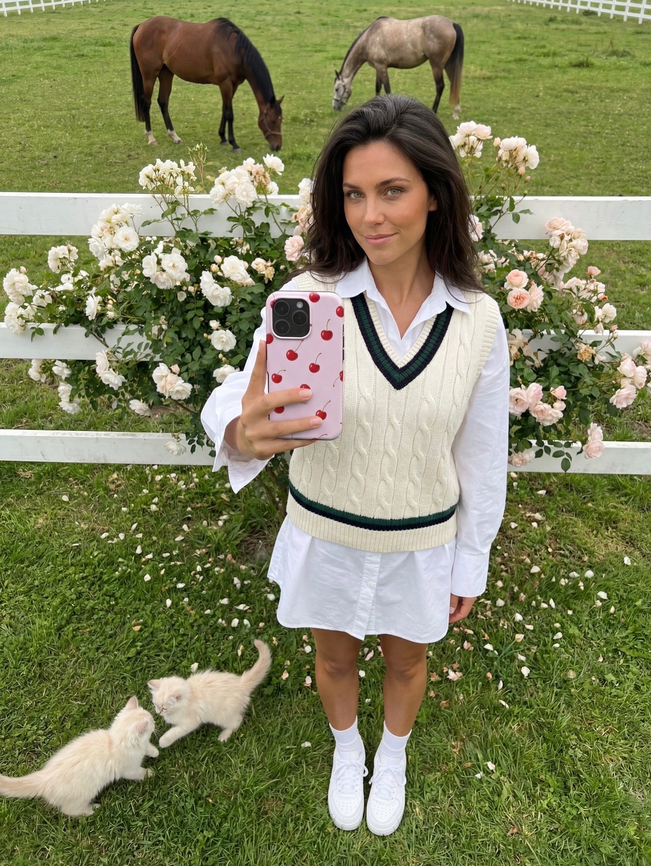 Trendy young woman outdoors holding phone with pink and red multicolored cherry patterned case, surrounded by white roses, green grass, and playful kittens.