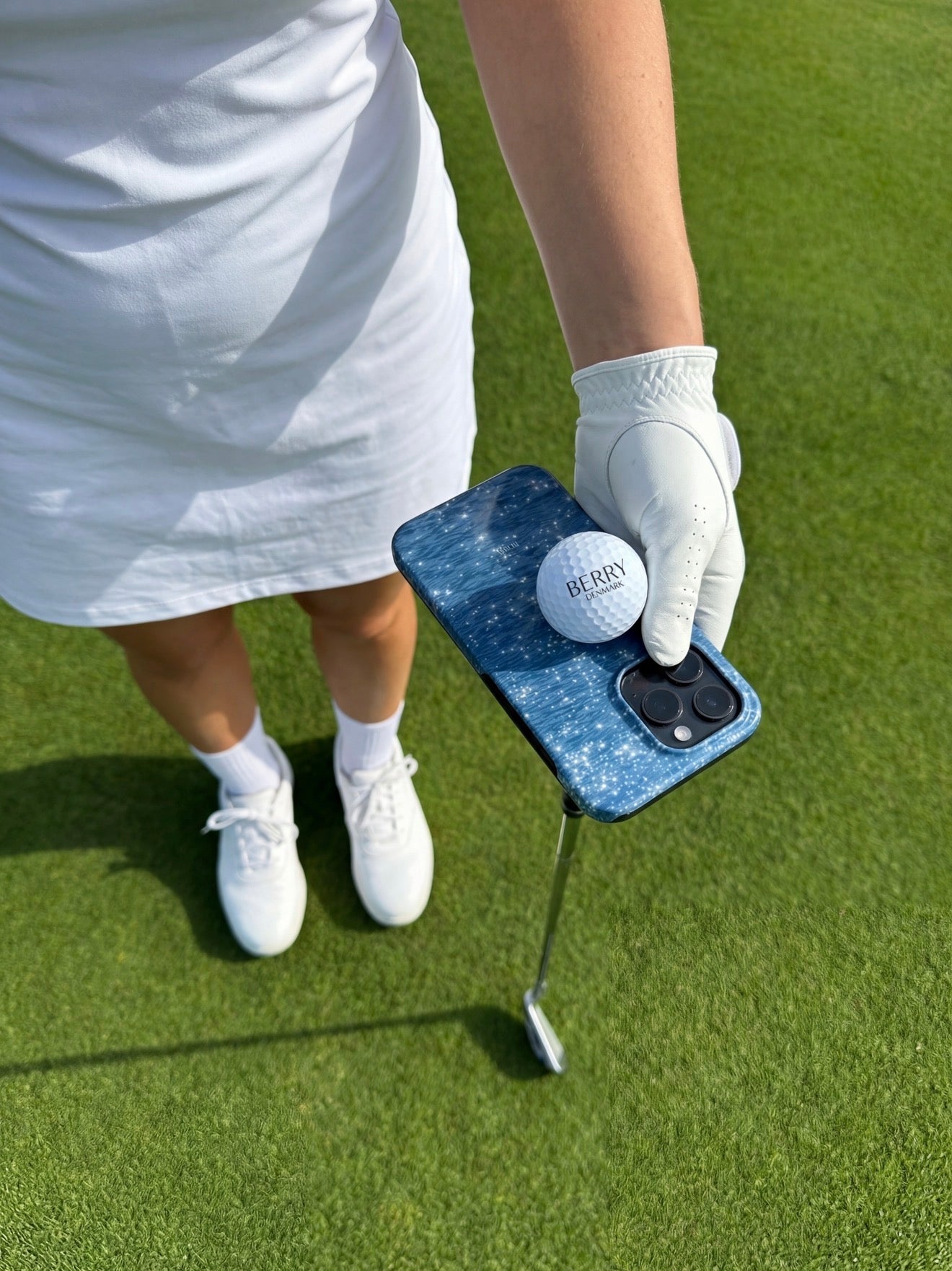 Trendy feminine hand in white glove holding a golf ball and blue multicolored phone case on a green golf course, styled for summer.
