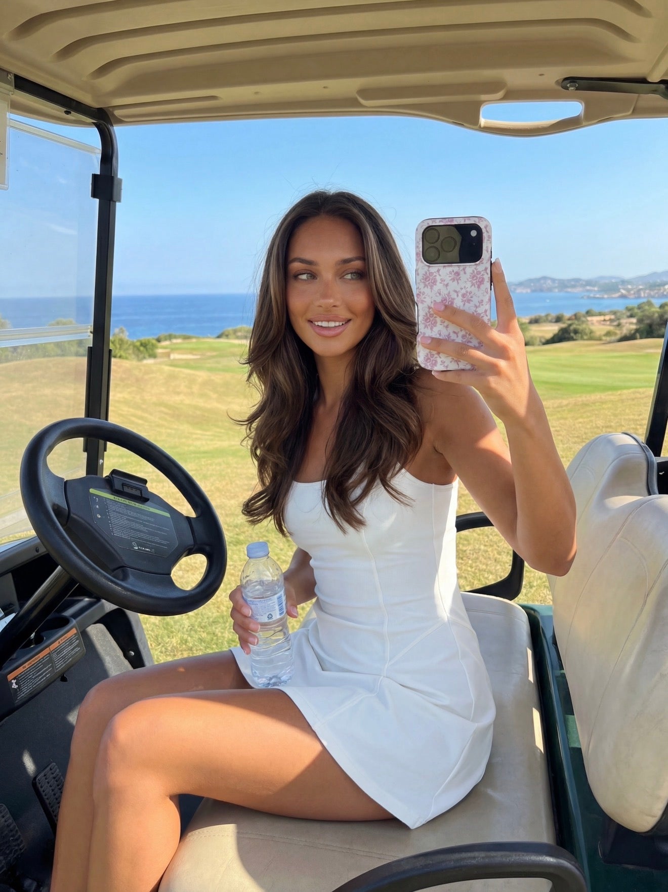 Trendy young woman with long hair wearing a white dress taking a selfie with a flerfarvet pink and white phone case inside a golf cart by the sea.