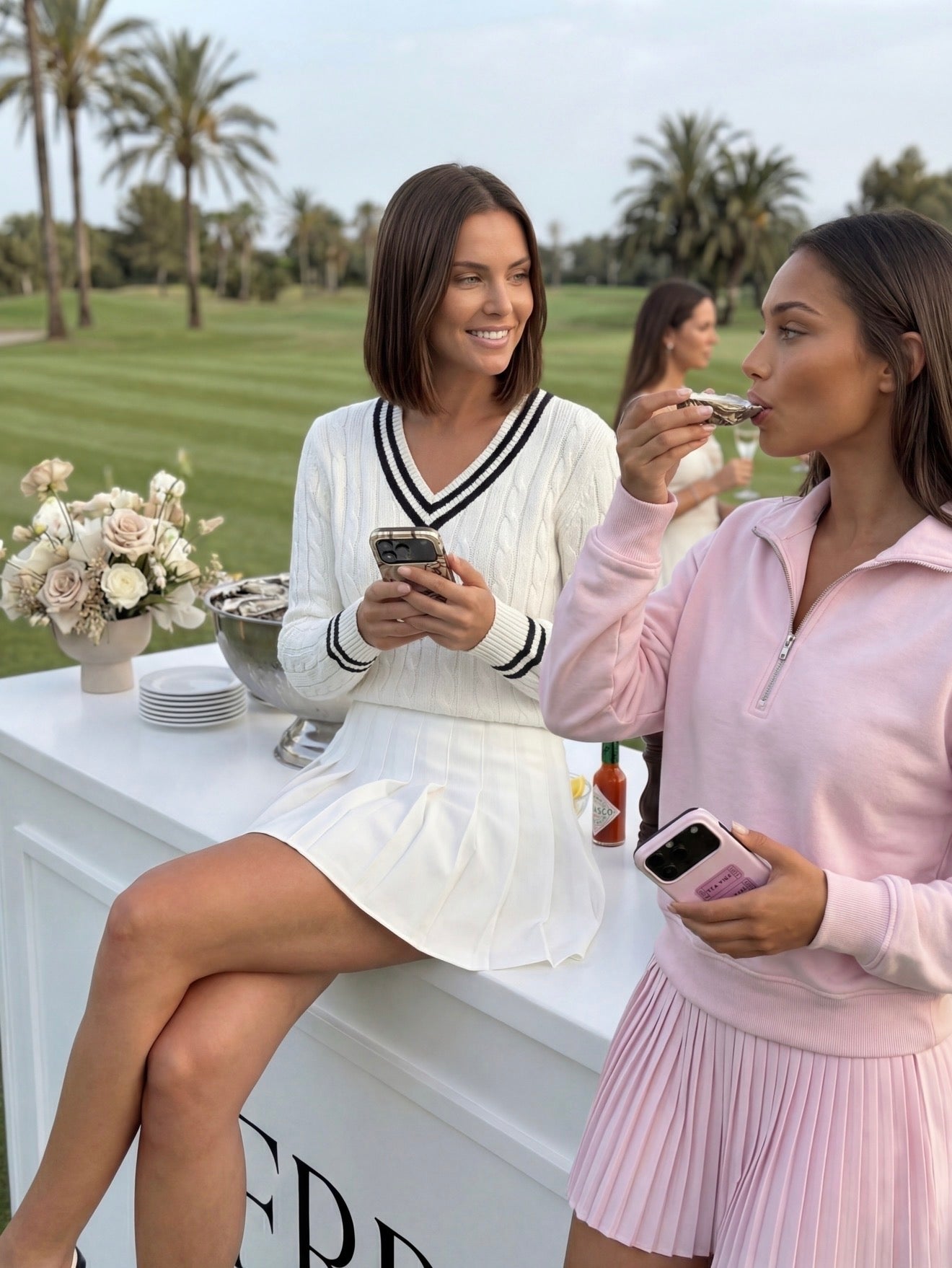 Two stylish women enjoying a garden tea time moment, one with a white pleated skirt and sweater, the other in a pink top and skirt, both holding phones.