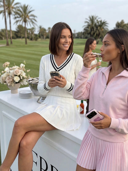 Two stylish women enjoying a garden tea time moment, one with a white pleated skirt and sweater, the other in a pink top and skirt, both holding phones.