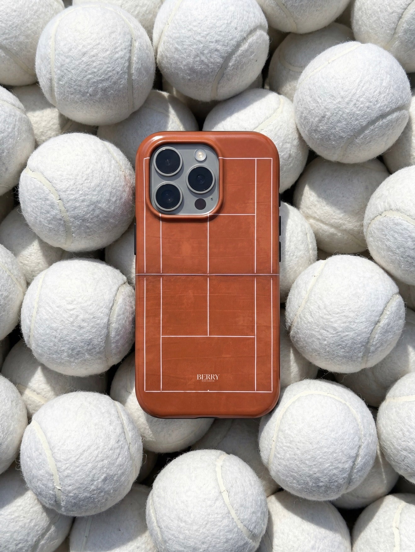 Trendy phone case in brown and orange with a tennis court design, placed among soft white tennis balls for a chic and sporty vibe.