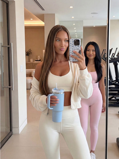 Two stylish women in trendy athleisure wear taking a mirror selfie, one holding a blue tumbler with a phone case featuring a multicolored pattern.