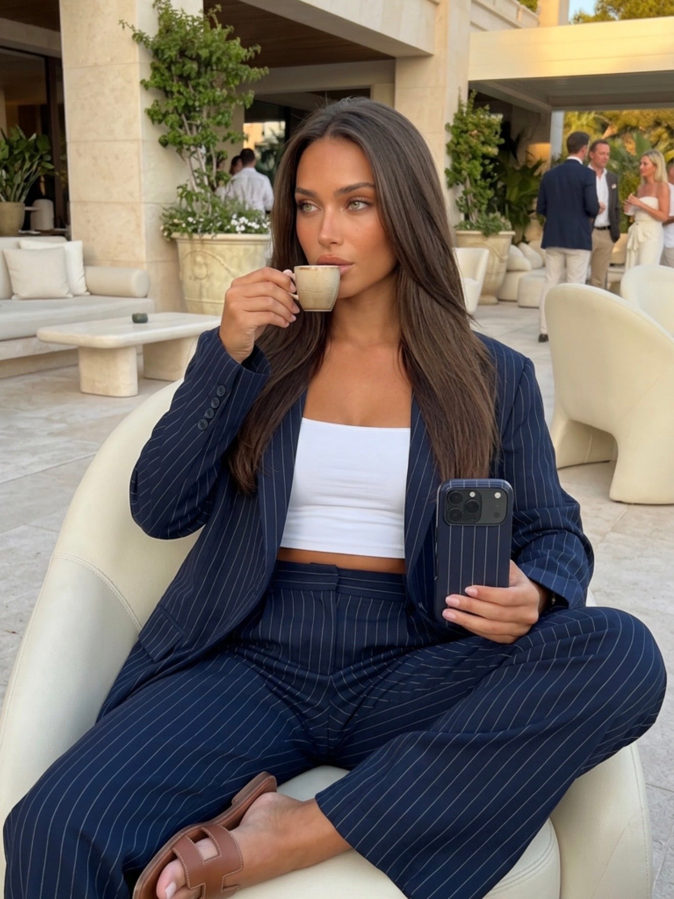 Trendy woman wearing a blue pinstripe suit with a white top, relaxing while sipping coffee and holding a matching phone case in an elegant outdoor setting.