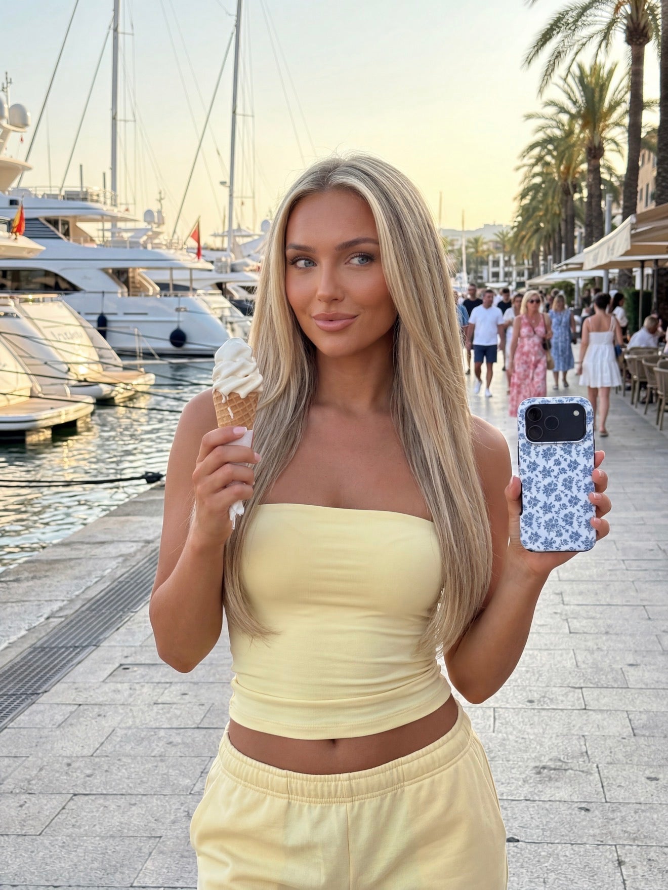Trendy young woman holding an ice cream cone and a phone with a multicolored white and blue floral patterned case by the harbor on a sunny day.