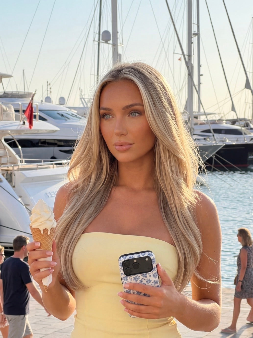 Trendy young woman with long blonde hair holding ice cream and a phone with a multicolored blue and white patterned case, standing near a marina with boats.