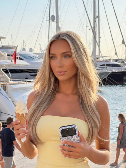 Trendy young woman with long blonde hair holding ice cream and a phone with a multicolored blue and white patterned case, standing near a marina with boats.