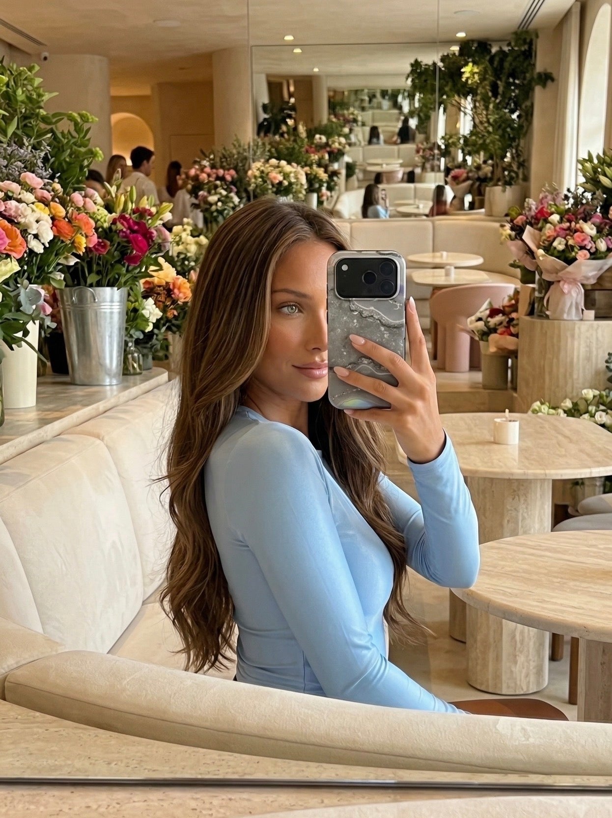 Trendy young woman with long brown hair taking a selfie with a chic silver marble quartz phone case in a stylish, flower-filled cafe setting.