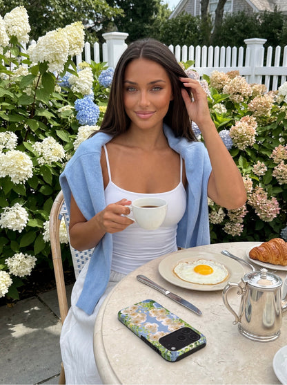 Trendy woman enjoying breakfast outdoors with coffee, wearing a light blue sweater and white top, natural floral phone case on the table beside croissant and sunny-side-up egg.