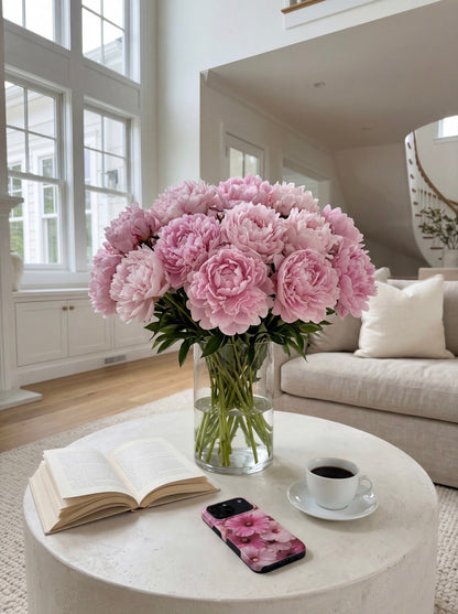 Aesthetic bouquet of pink and red multicolored flowers sits on a white table near a trendy phone case, open book, and coffee cup in a bright modern living room.