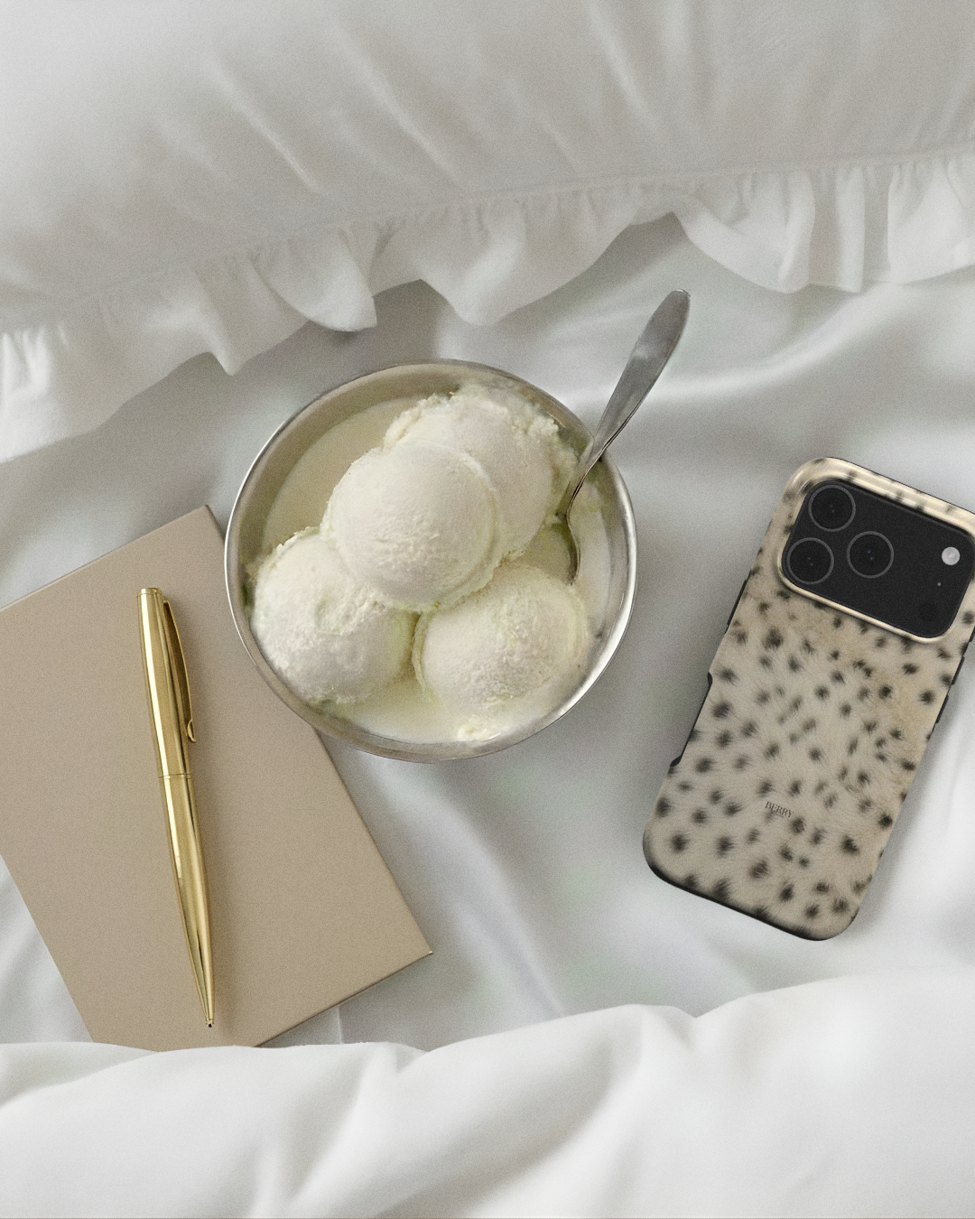 Trendy phone case with gold and bronze spotted pattern beside a gold pen and notebook on soft white fabric next to a bowl of creamy ice cream scoops.