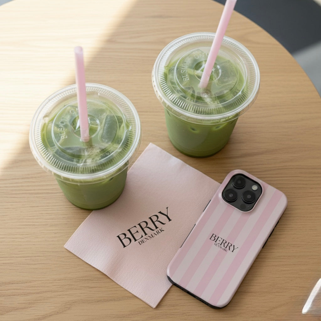 Pink striped phone case with feminine design placed on a wooden table next to two iced green drinks and a Berry Denmark napkin, trendy and stylish aesthetic.
