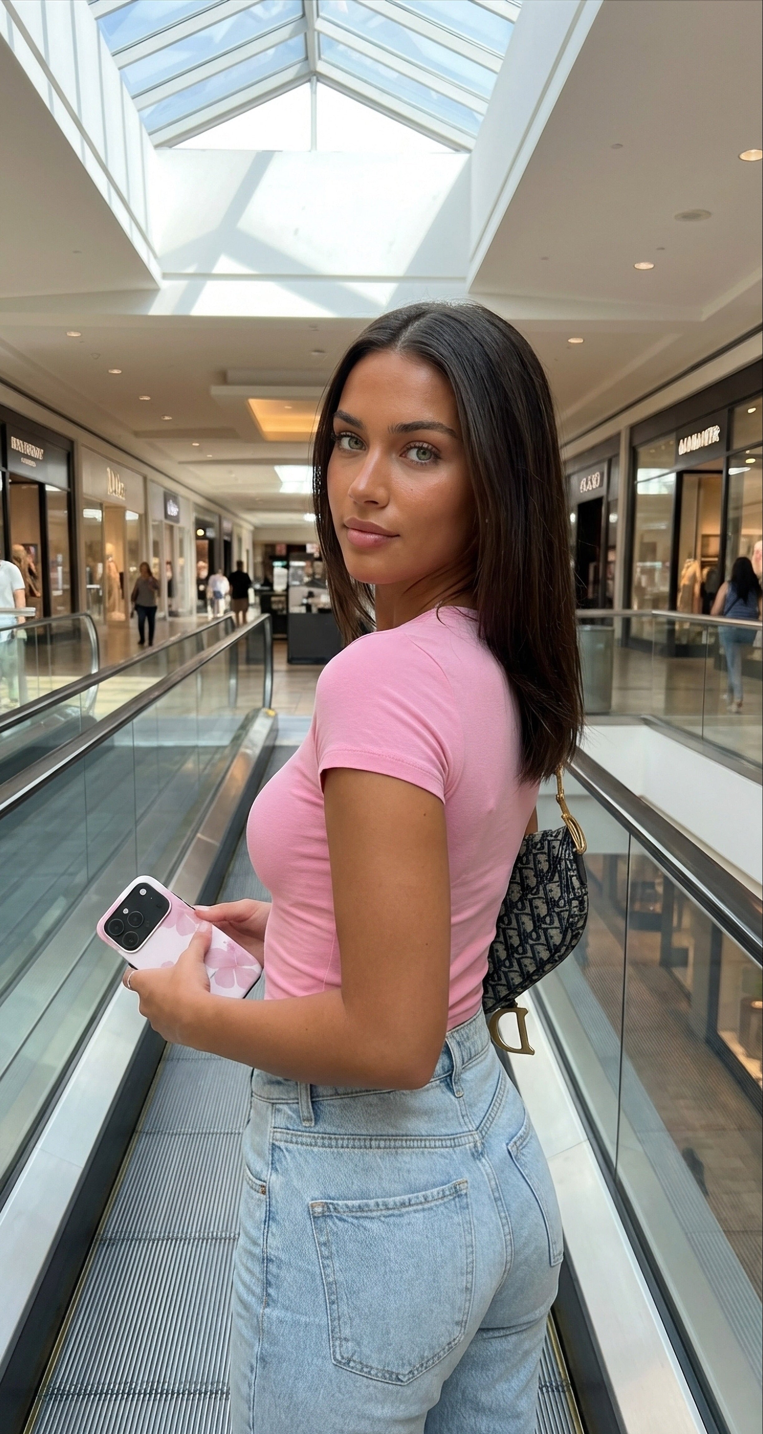 Trendy young woman in a soft pink top holding a phone with a floral soft pink case, standing on an escalator in a bright shopping mall.
