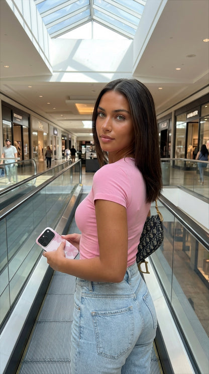 Trendy young woman in a soft pink top holding a phone with a floral soft pink case, standing on an escalator in a bright shopping mall.