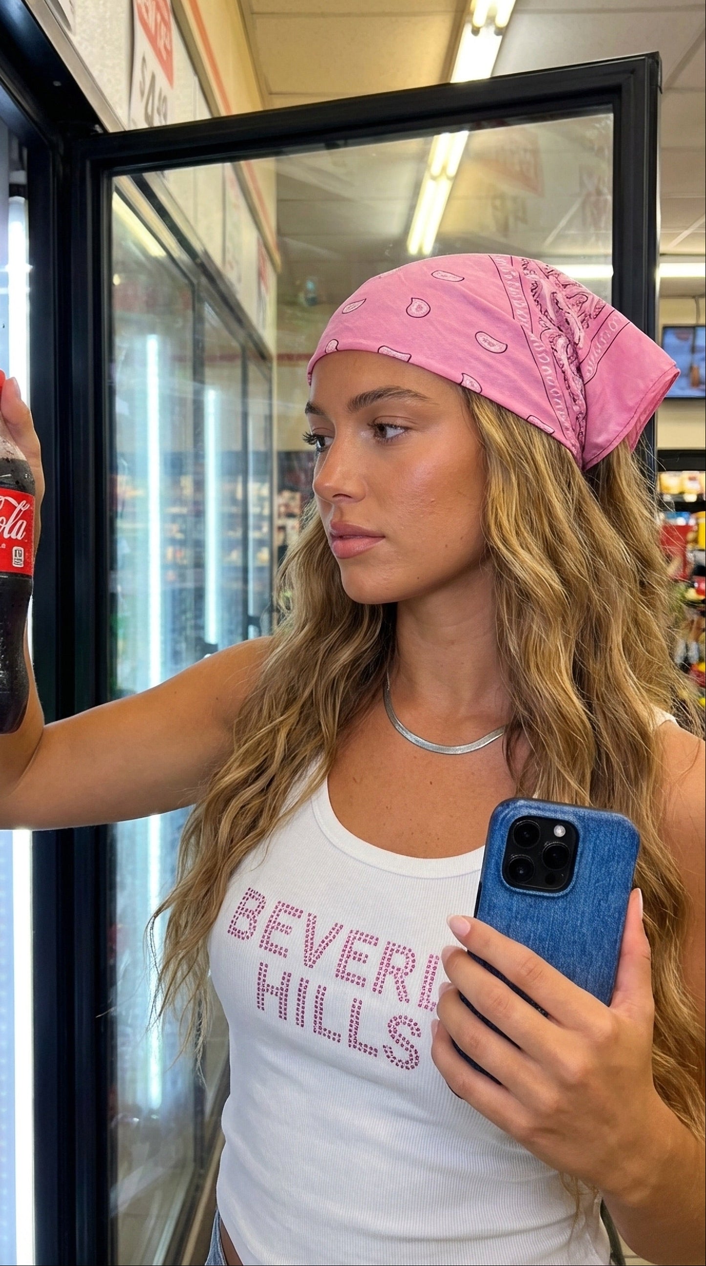 Young woman with wavy hair and pink bandana holding a phone with blue jeans-style case, looking at a cold drink inside a store refrigerator.
