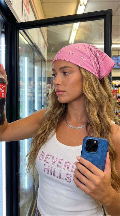 Young woman with wavy hair and pink bandana holding a phone with blue jeans-style case, looking at a cold drink inside a store refrigerator.