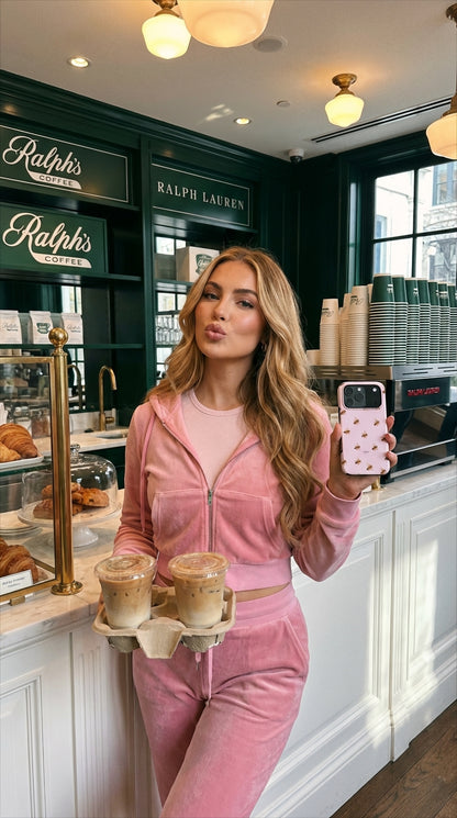 Trendy woman in pink velour tracksuit holding a pink patterned phone case and iced coffee drinks in a stylish café setting with natural light.