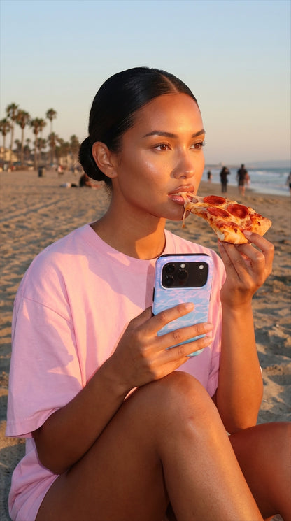 Trendy young woman on the beach holding a blue and purple multicolored phone case while enjoying a slice of pepperoni pizza at sunset.