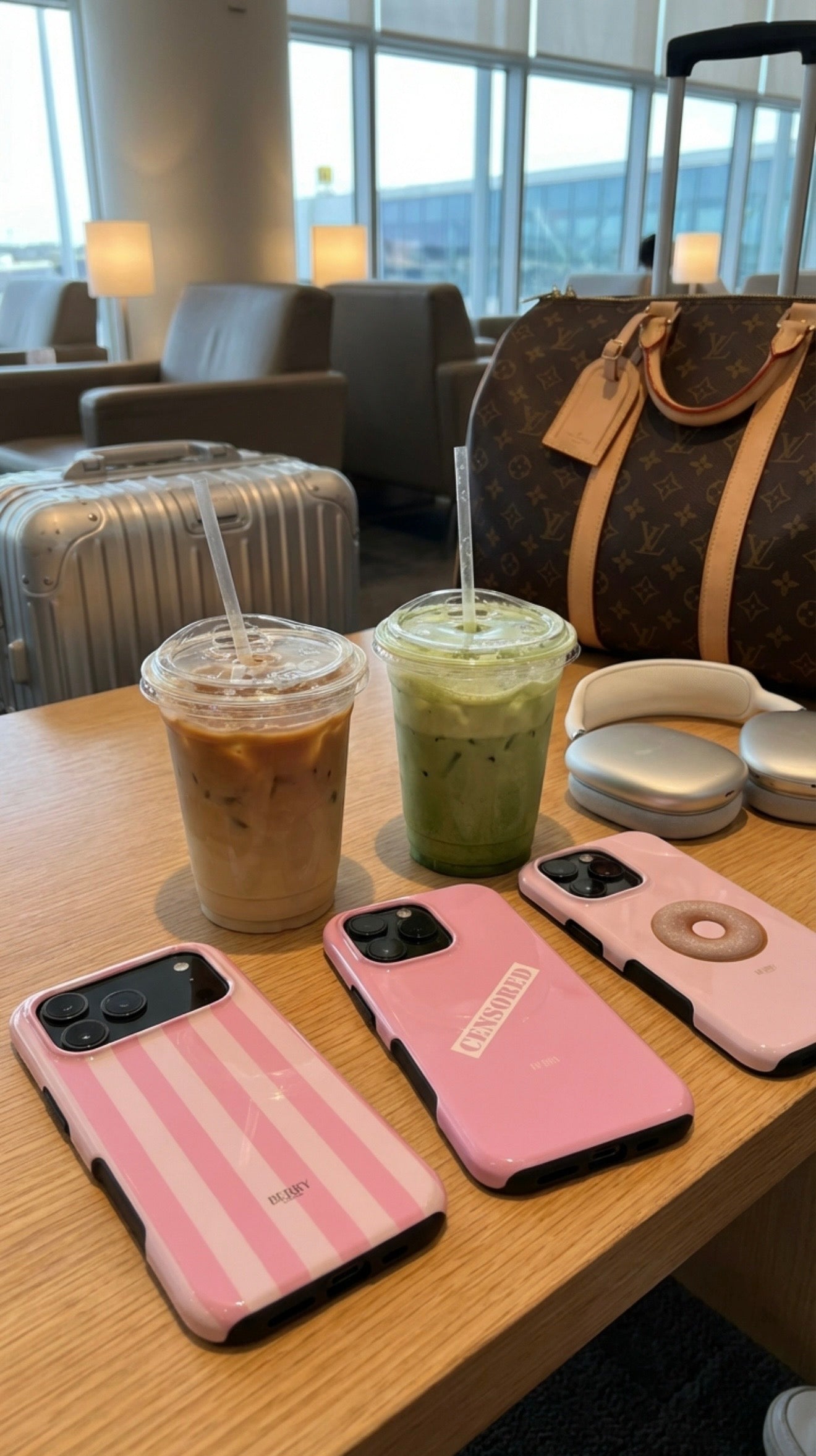 Three trendy pink phone cases with iced drinks on a wooden table in a bright lounge, featuring a designer brown bag and silver headphones nearby.