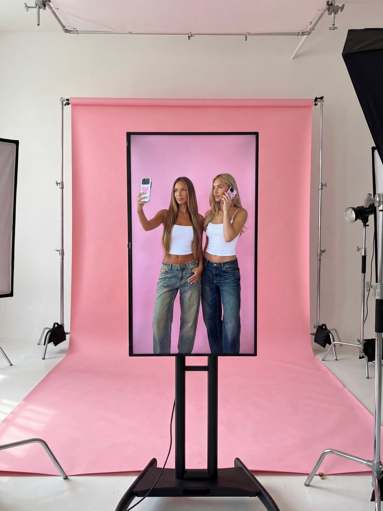 Two stylish women in white tops and jeans posing for trendy phone photos against a pink backdrop, showcasing feminine and popular aesthetic vibes.