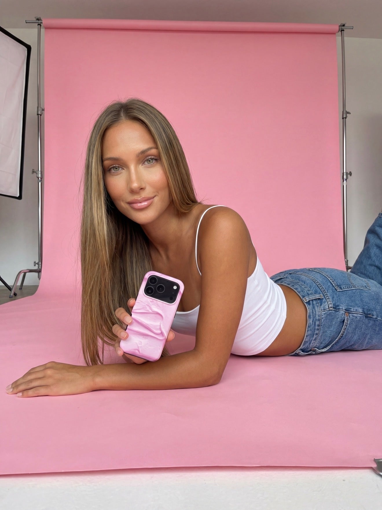Trendy young woman lying on pink backdrop holding a pink and flerfarvet phone case with a feminine and stylish design.